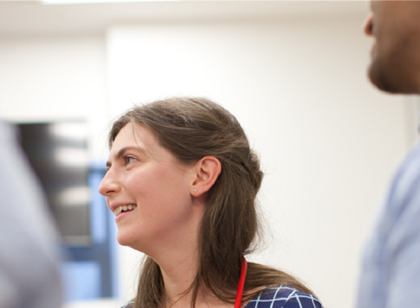 A photo of a young female doctor's face, with a stethoscope just visible around her neck. She is turned to the left, towards an out-of-focus person, with another out-of-focus man on the right of the image.