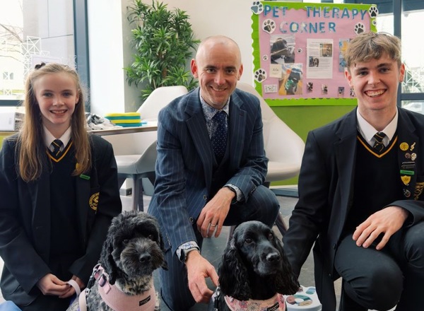 A photo of brother and sister pupils Paul and Margot Kennedy and their headteacher Huw Brophy from St Thomas More RC College in Denton visiting the proton beam therapy centre at The Christie to meet resident therapy dogs Lilo and Luna.