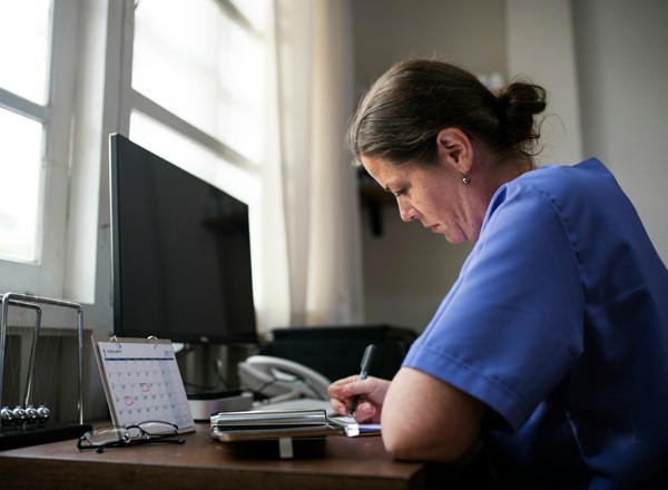 A photo of a woman taking notes at a desk with a computer and a calendar on it.