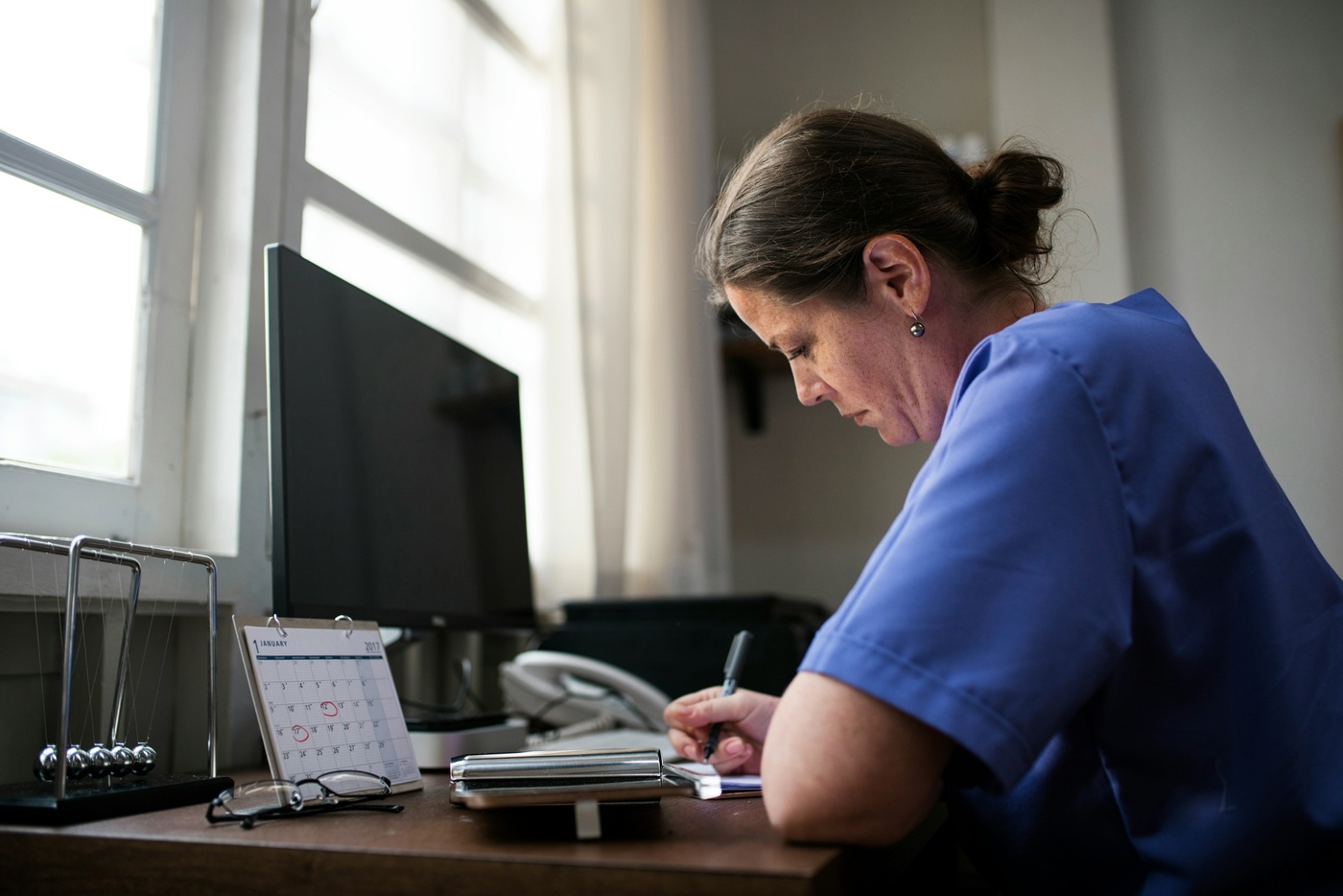 A photo of a woman taking notes at a desk with a computer and a calendar on it.