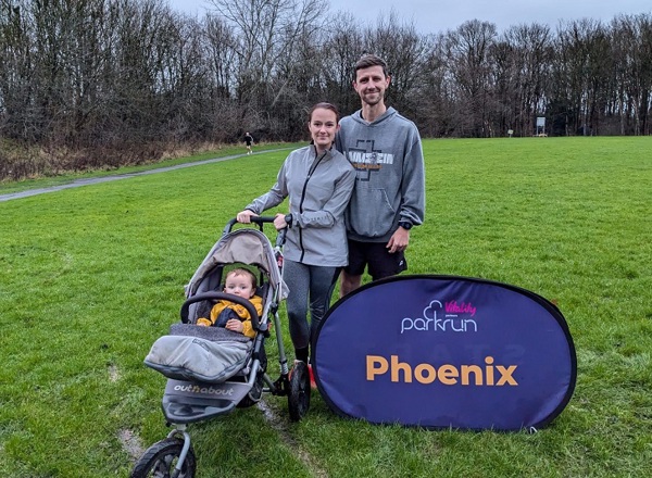 A photo of Christie patient Zac Hinchcliffe and his wife Danni in running clothes, with their baby Fenn in a push chair. The 3 are in a park at a parkrun event, a sign reads 'parkrun Phoenix'.