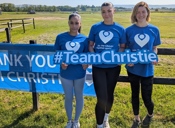 A photo of Christie nurses Rasleen Sharma, Rhiana Smith and Abbie Gunton wearing Christie Charity t-shirts and holding a sign that reads '#TeamChristie'; they are standing in front of a field.