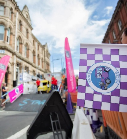 A photo of the Totally Stockport Krazy Races soapbox derby event, with a purple starting flag in the foreground and a blurred yellow soapbox racer in the back.