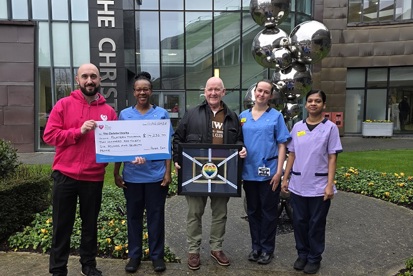 A photo of Christie fundraiser Peter Day standing in front of The Christie's main entrance with 3 members of staff and Christie Charity staff member Adam George. peter is holding a completed framed Christie Challenge medal and Adam is holding a big cheque from Peter to The Christie Charity for £14,236.