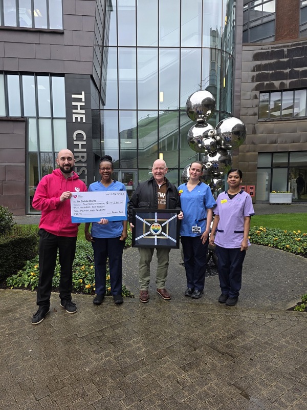 A photo of Christie fundraiser Peter Day standing in front of The Christie's main entrance with 3 members of staff and Christie Charity staff member Adam George. peter is holding a completed framed Christie Challenge medal and Adam is holding a big cheque from Peter to The Christie Charity for £14,236.