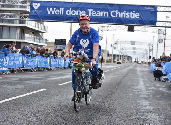 A photo of Tony O'Dwyer riding a bike along the Blackpool promenade with a Basil Brush toy on the front of his bike; a sign above him reads "Well done Team Christie".