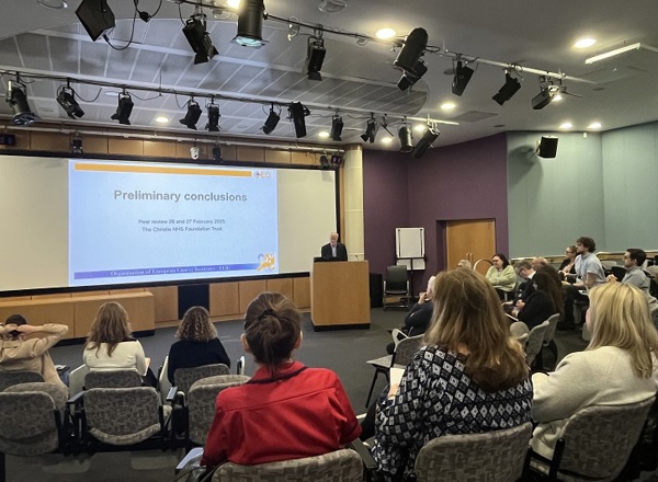 A photo of Simon Osbert from the OECI presenting to a group of people in The Christie auditorium; the screen read 'Preliminary conclusions: Peer review 26 and 27 February 2025, The Christie NHS Foundation Trust'.