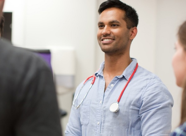 A photo of a young doctor with a stethoscope around his neck. He is looking to the left of the image, where 2 other people can be seen out of focus, with a woman on the right-hand side of the image also visible.