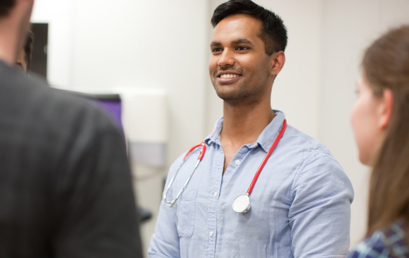 A photo of a young doctor with a stethoscope around his neck. He is looking to the left of the image, where 2 other people can be seen out of focus, with a woman on the right-hand side of the image also visible.
