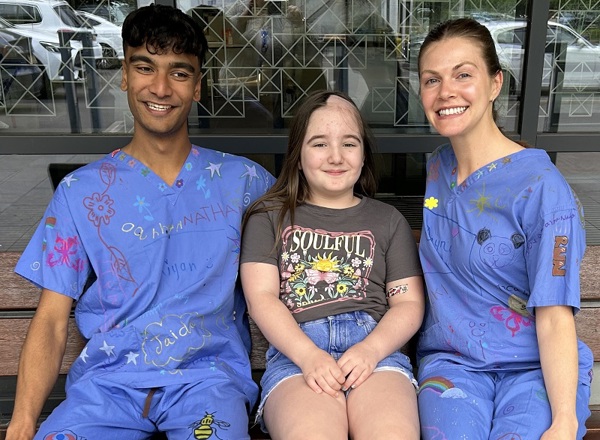 A photo of Christie radiographers Kiyan and Dayna wearing their decorated scrubs and sitting on a bench with young Christie patient Esmai Evans.