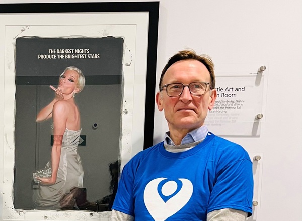 A photo of Dr Sacha Howell standing outside The Christie art room in front of photo of Sarah Harding which reads 'The darkest nights produce the brightest stars'.