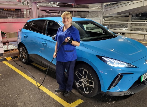 A photo of Christie at Home nurse Sarah Richardson standing next to a blue electric MG4 car.