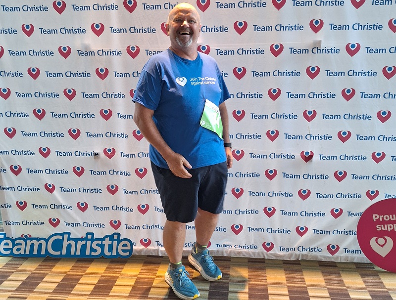 A photo of Mark Love, Christie fundraiser, wearing a Christie Charity t-shirt and standing in front of a Team Christie background.