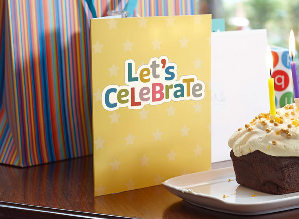 A photo of a birthday cake with lit candles, a glass of orange juice and a card reading 'Let's celebrate'.