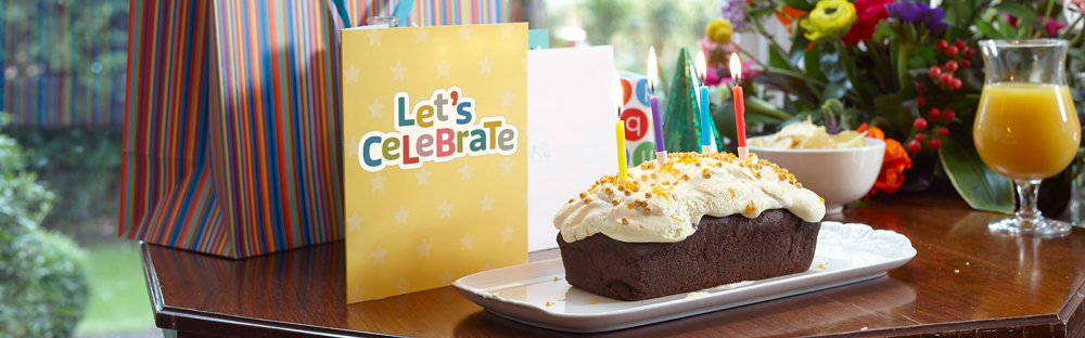 A photo of a birthday cake with lit candles, a glass of orange juice and a card reading 'Let's celebrate'.