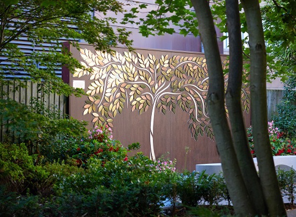 A photo of the Tree of Hope at The Christie, the shape of a tree made up of metal leaves with people's names on them on a brown fence. 