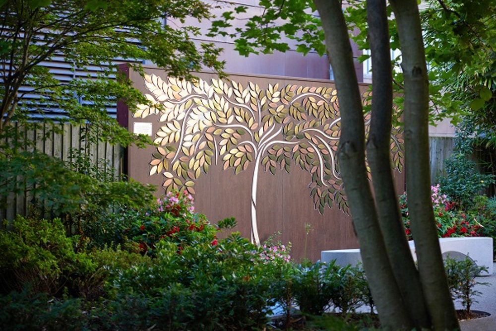 A photo of the Tree of Hope at The Christie, the shape of a tree made up of metal leaves with people's names on them on a brown fence. 