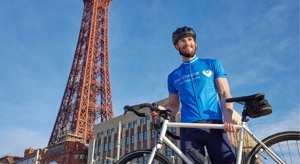 A photo of a male cyclist holding a bike and standing in front of the Blackpool tower.