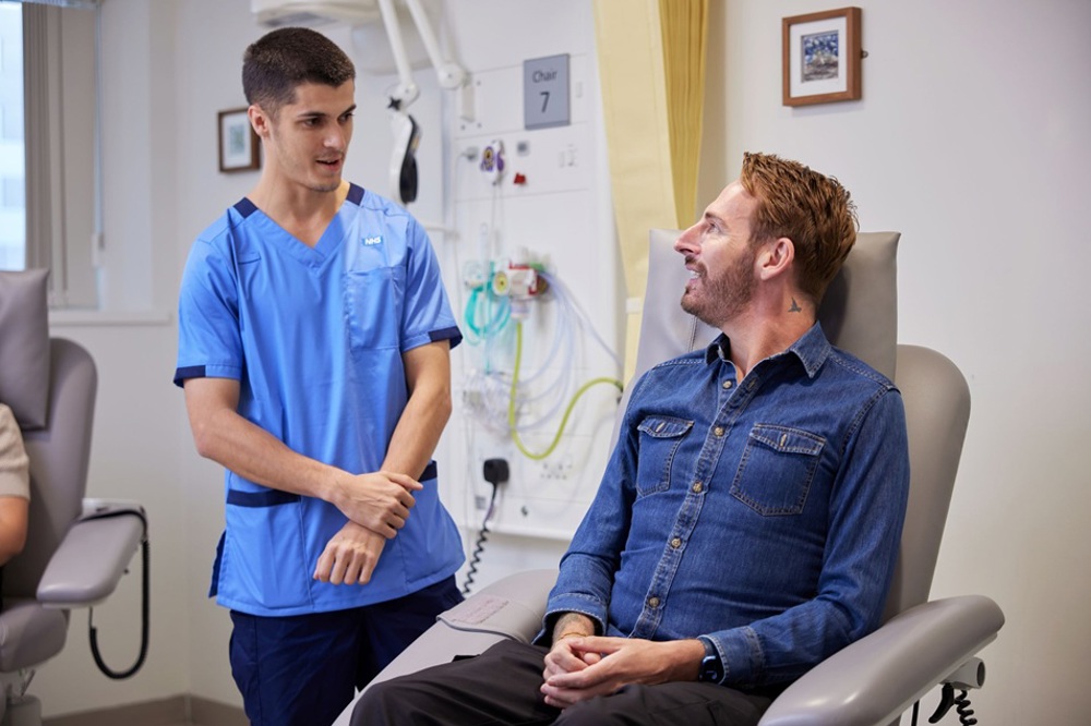 A photo of a man sitting in a chemotherapy treatment chair accompanied by a male staff member in a clinical trials uniform.