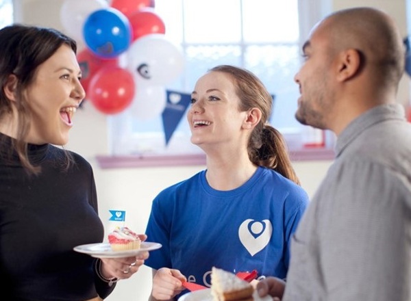 A photo of a fundraising event for The Christie Charity. One female fundraiser in the middle of the photo is wearing a blue Christie Charity t-shirt and she has another female fundraiser on one side and a male fundraiser on the other side. They are holding cake, and balloons and bunting can be seen in the background.