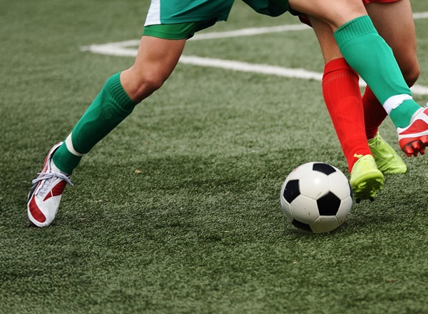 A photo of 2 sets of feet kicking a football; one wearing green socks and the other wearing red socks.