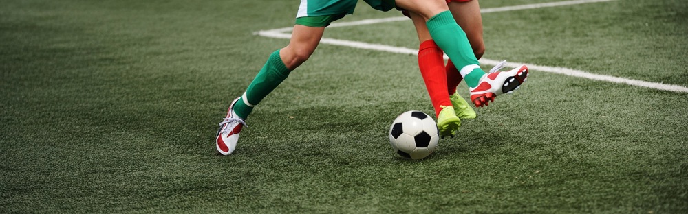 A photo of 2 sets of feet kicking a football; one wearing green socks and the other wearing red socks.