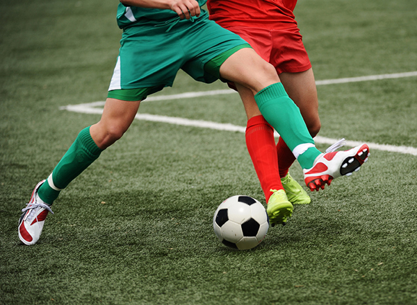 A photo of 2 sets of feet kicking a football; one wearing green socks and the other wearing red socks.