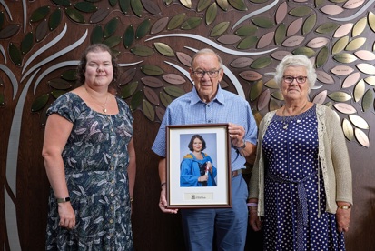 A photo of Jayne, Norman and Pearl Howell standing in front of The Christie Charity's Tree of Hope and holding a graduation photo of Claire.