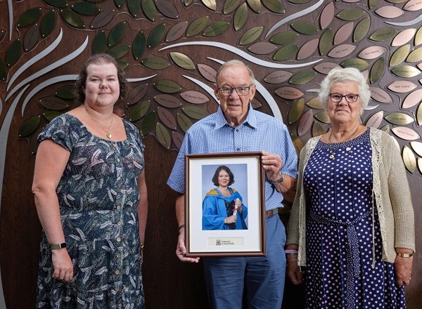 A photo of Jayne, Norman and Pearl Howell standing in front of The Christie Charity's Tree of Hope and holding a graduation photo of Claire.