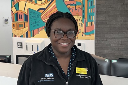 A photo of Aanu Majaro, receptionist at The Christie, standing in front of a mural that reads 'The Christie at Salford Royal'.