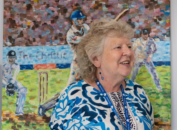 A photo of Pat Mountford, the artist in residence at The Christie, standing in front of a painting by a Christie patient.