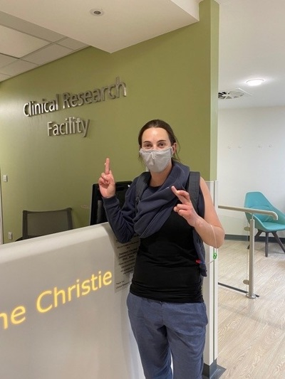 A photo of Christie patient Bex Hind wearing a surgical face mask and standing at the reception of the Clinical Research Facility at The Christie.