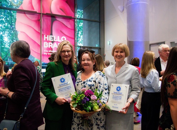 A photo of Tina Karabatsou, another woman and Catherine McBain. Tina and Catherine are holding plaques reading 'Tessa Jowell Centre of Excellence, Salford Royal Hospital, Northern Care Alliance', and the middle woman is holding a bunch of flowers.