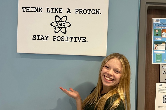 A photo of Eve Wilson, a former patient at the proton beam therapy centre at The Christie, stands in front of a sign that reads 'Think like a proton. Stay positive."
