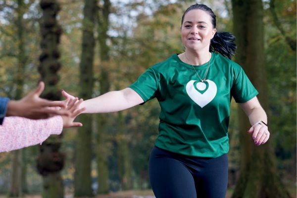 A photo of a woman running through a forest wearing a green Christie Charity t-shirt.
